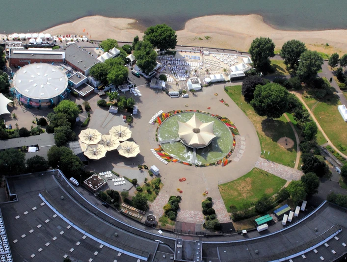Tanzbrunnen Köln mit Rheinterrassen und km 689 Cologne Beach Club, Luftaufnahme Das Bild zeigt eine Veranstaltungslocation am Rhein mit einem sternförmigen Pavillon und umliegenden Grünflächen.The picture shows an event location on the Rhine with a star-shaped pavilion and surrounding green areas.