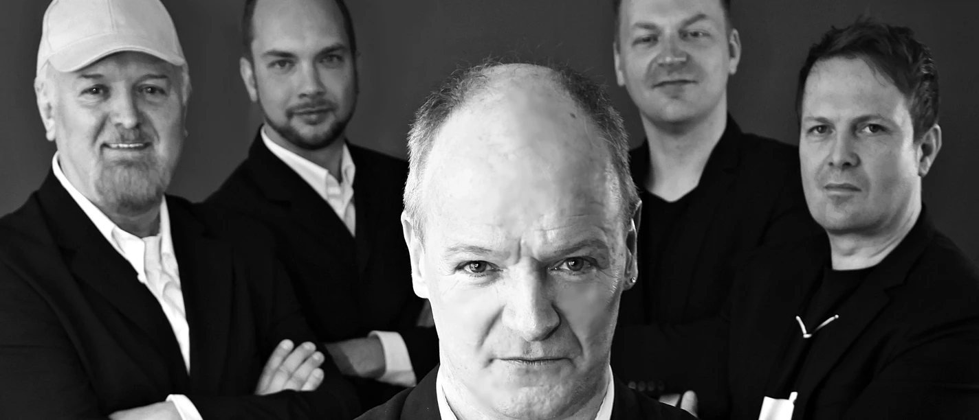 Pressefoto Pick Up-2.jpg Fünf Männer in Anzügen vor dunklem Hintergrund, einer steht vorn, die anderen dahinter mit verschränkten Armen.