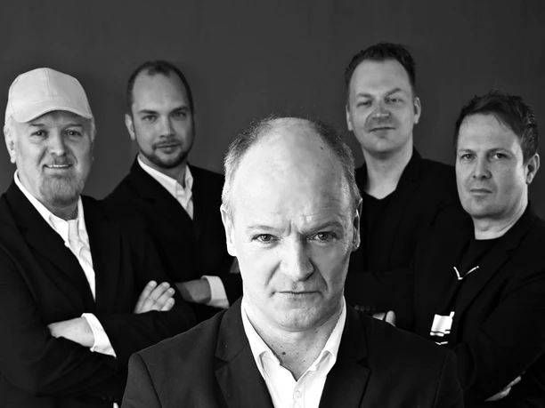 Pressefoto Pick Up-2.jpg Fünf Männer in Anzügen vor dunklem Hintergrund, einer steht vorn, die anderen dahinter mit verschränkten Armen.