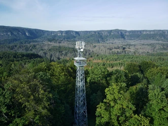 Janov uitzichtpunt Een zendmast voor mobiele telefonie rijst op uit een dicht, groen bos, met beboste heuvels en een heldere hemel op de achtergrond.