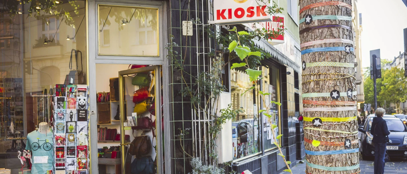 Belgisches Viertel in Köln Ein traditioneller Kiosk in Köln bietet farbenfrohe Waren aller Art. Ein Baum vor dem Laden ist mit bunten Bändern und Sternen dekoriert, was der Straße ein lebendiges Flair verleiht.