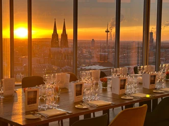 Firmenfeier_KölnSKY Köln, Domblick bei Sonnenuntergang durch Fenster, gedeckte Restauranttische vorne.
