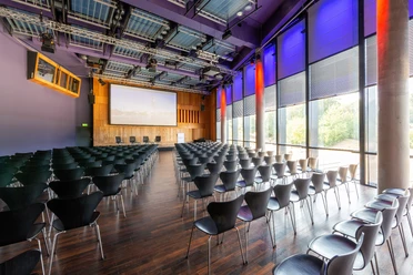 KOMED-Saal Moderner Konferenzraum mit dunklen Stühlen, großem Bildschirm und lila beleuchteten Wänden.</p>