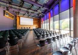 KOMED-Saal Moderner Konferenzraum mit dunklen Stühlen, großem Bildschirm und lila beleuchteten Wänden.</p>Modern conference room with dark chairs, large screen and purple-lit walls.</p> <p