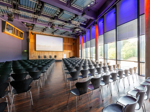 KOMED-Saal Moderner Konferenzraum mit dunklen Stühlen, großem Bildschirm und lila beleuchteten Wänden.</p>