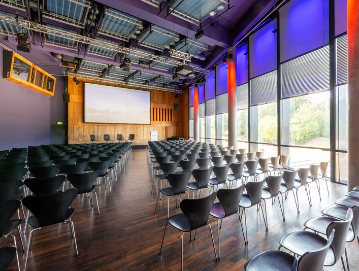 KOMED-Saal Moderner Konferenzraum mit dunklen Stühlen, großem Bildschirm und lila beleuchteten Wänden.</p>Modern conference room with dark chairs, large screen and purple-lit walls.</p> <p