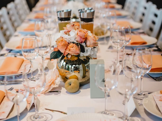 4711 Duftmenü Tisch Tisch mit weißer Tischdecke, dekoriert mit orangefarbenen Servietten und Blumenarrangements.