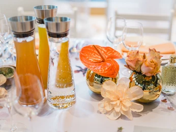 4711 Duftmenü Tisch 2 Blumendekorierter Tisch mit Glasflaschen und zwei goldenen Vasen, gefüllt mit Blumenarrangements.Flower-decorated table with glass bottles and two golden vases filled with flower arrangements.