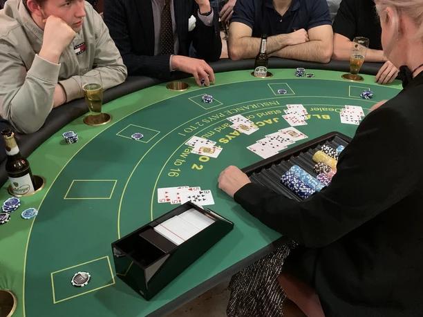 Black Jack 01 Poker players at a round green table with cards, drinks and chips in a convivial atmosphere.