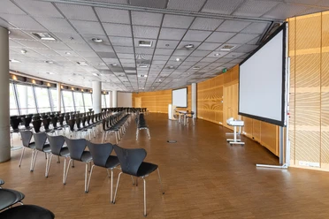 Raum 1-3 Ein moderner Konferenzraum mit leeren Stuhlreihen und zwei großen Bildschirmen.</p>