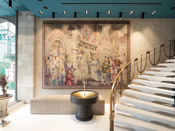 Dufthaus Interior 1 Tapestry in a historic building, showing a lively street scene; elegant staircase in the foreground.
