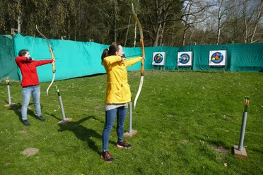 Bogenschießen.JPG Zwei junge Frauen stehen auf einer grünen Wiese und schießen mit dem Bogen auf Zielscheiben. Die Bögen sind gespannt und schussbereit.