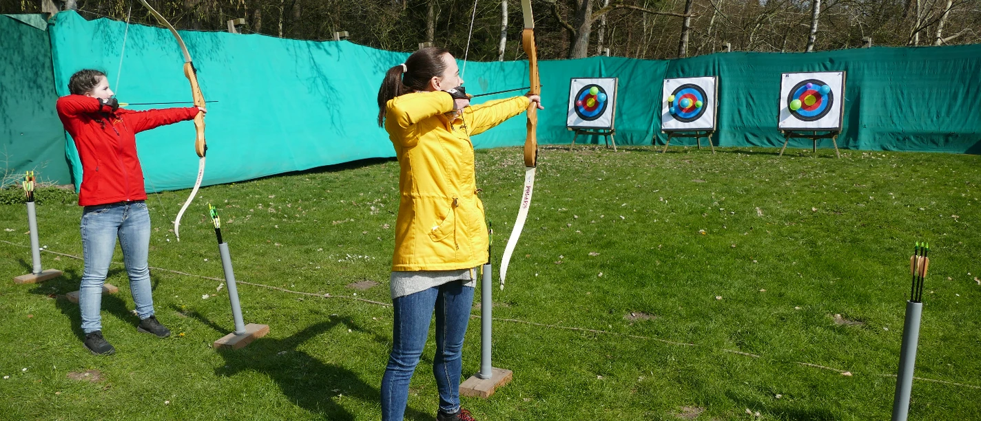 Bogenschießen.JPG Zwei junge Frauen stehen auf einer grünen Wiese und schießen mit dem Bogen auf Zielscheiben. Die Bögen sind gespannt und schussbereit.