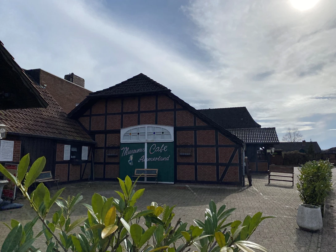 Bild 4.jpg Das Bild zeigt ein traditionelles Fachwerkgebäude mit der Aufschrift "Mühlen Café Ammerland". Der Vordergrund sind Pflanzen zu sehen, die dem Bild eine natürliche Note geben.