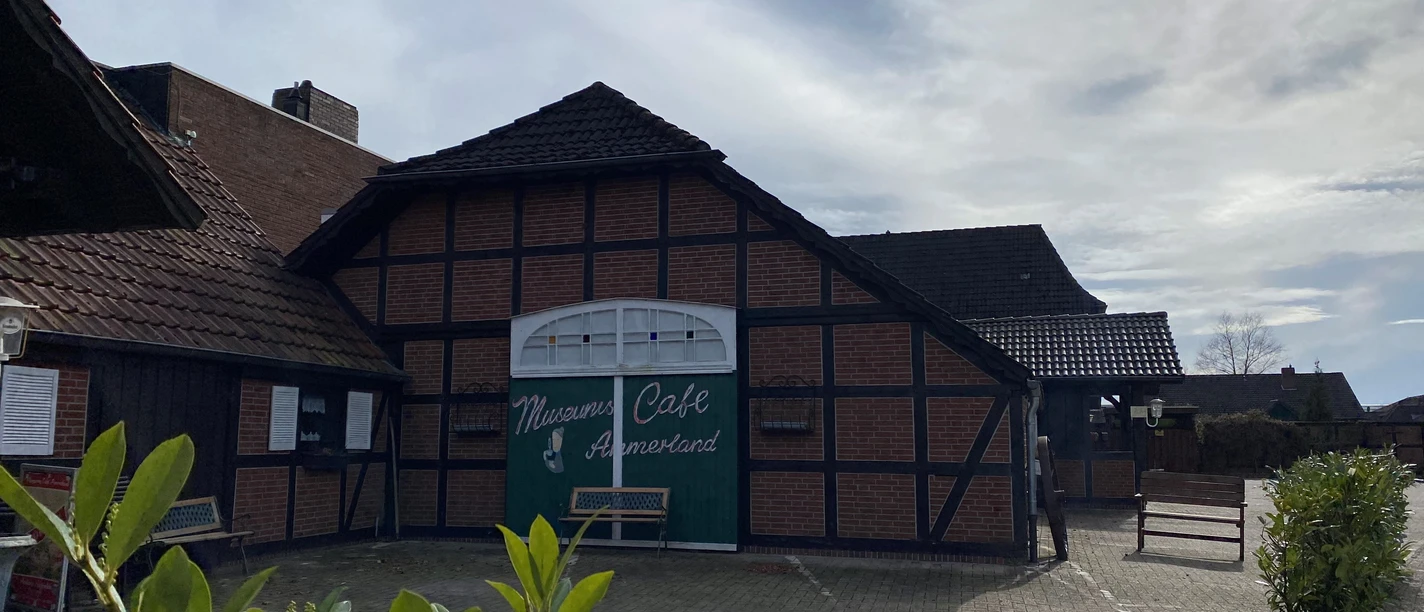 Bild 4.jpg Das Bild zeigt ein traditionelles Fachwerkgebäude mit der Aufschrift "Mühlen Café Ammerland". Der Vordergrund sind Pflanzen zu sehen, die dem Bild eine natürliche Note geben.