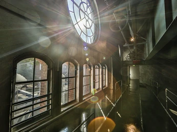E-Werk Köln Ambiente Industriehalle mit großen Rundbogenfenstern, Sonnenstrahlen durchfluten den Raum, Treppe im Hintergrund.</p>Industrial hall with large arched windows, sunlight flooding the room, staircase in the background.</p> <p