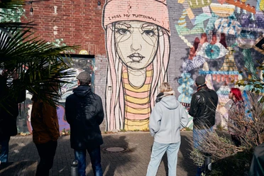 Ehrenfeld Street Art Tour A group of people is looking at a large street art painting on a brick wall, depicting a melancholic young woman wearing a beanie. Plants are visible in the foreground.