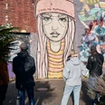 Ehrenfeld Street Art Tour Eine Gruppe von Menschen betrachtet ein großes Street-Art-Gemälde auf einer Backsteinmauer, das eine melancholische, junge Frau in Mütze zeigt. Im Vordergrund sind Pflanzen sichtbar.A group of people is looking at a large street art painting on a brick wall, depicting a melancholic young woman wearing a beanie. Plants are visible in the foreground.