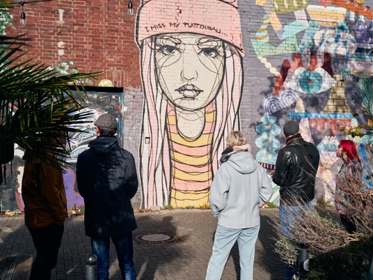 Ehrenfeld Street Art Tour Eine Gruppe von Menschen betrachtet ein großes Street-Art-Gemälde auf einer Backsteinmauer, das eine melancholische, junge Frau in Mütze zeigt. Im Vordergrund sind Pflanzen sichtbar.A group of people is looking at a large street art painting on a brick wall, depicting a melancholic young woman wearing a beanie. Plants are visible in the foreground.