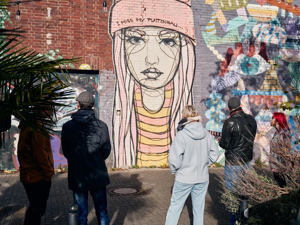 Ehrenfeld Street Art Tour Eine Gruppe von Menschen betrachtet ein großes Street-Art-Gemälde auf einer Backsteinmauer, das eine melancholische, junge Frau in Mütze zeigt. Im Vordergrund sind Pflanzen sichtbar.