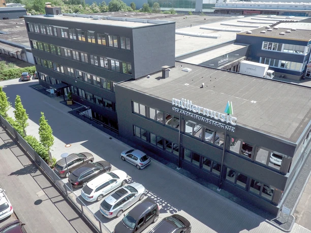 Firmensitz Modern office building with "Müllermusic" lettering on the façade, surrounded by parking spaces.</p> <p