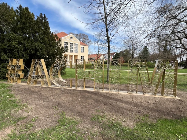 Achim-Schriftzug Großer #Achim-Schriftzug aus Metall im Grünen, neben kahlen Bäumen und einem rosafarbenen Gebäude.