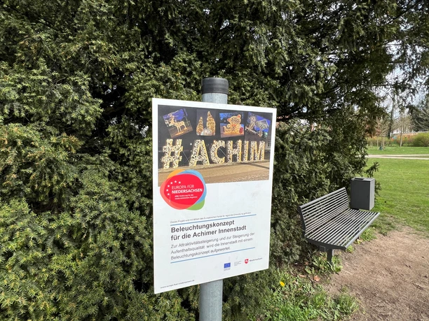 Achim-Schriftzug Schild Schild mit der Aufschrift "#ACHIM" vor Büschen in einem Park, informiert über Beleuchtungskonzept.