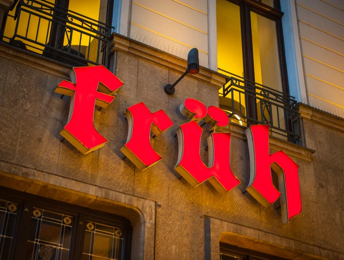 Brauhaus FRÜH am Dom Leuchtendes, rotes "Früh"-Schild an historischer Gebäudefassade bei warmem Abendlicht.</p>Bright red "Früh" sign on historic building façade in warm evening light</p>.