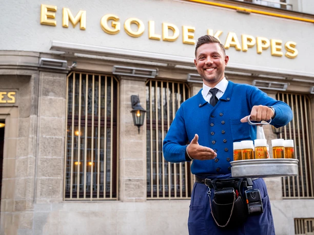 Brauhaus FRÜH "Em Golde Kappes" Kellner in blauer Jacke serviert frische Biere vor Gaststätte "Em Golde Kappes" mit historischer Fassade.