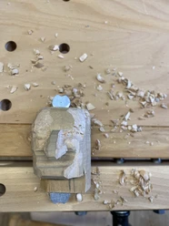 Houten sculptuur van een gezicht aan het werk op een werkbank, omringd door houtkrullen.