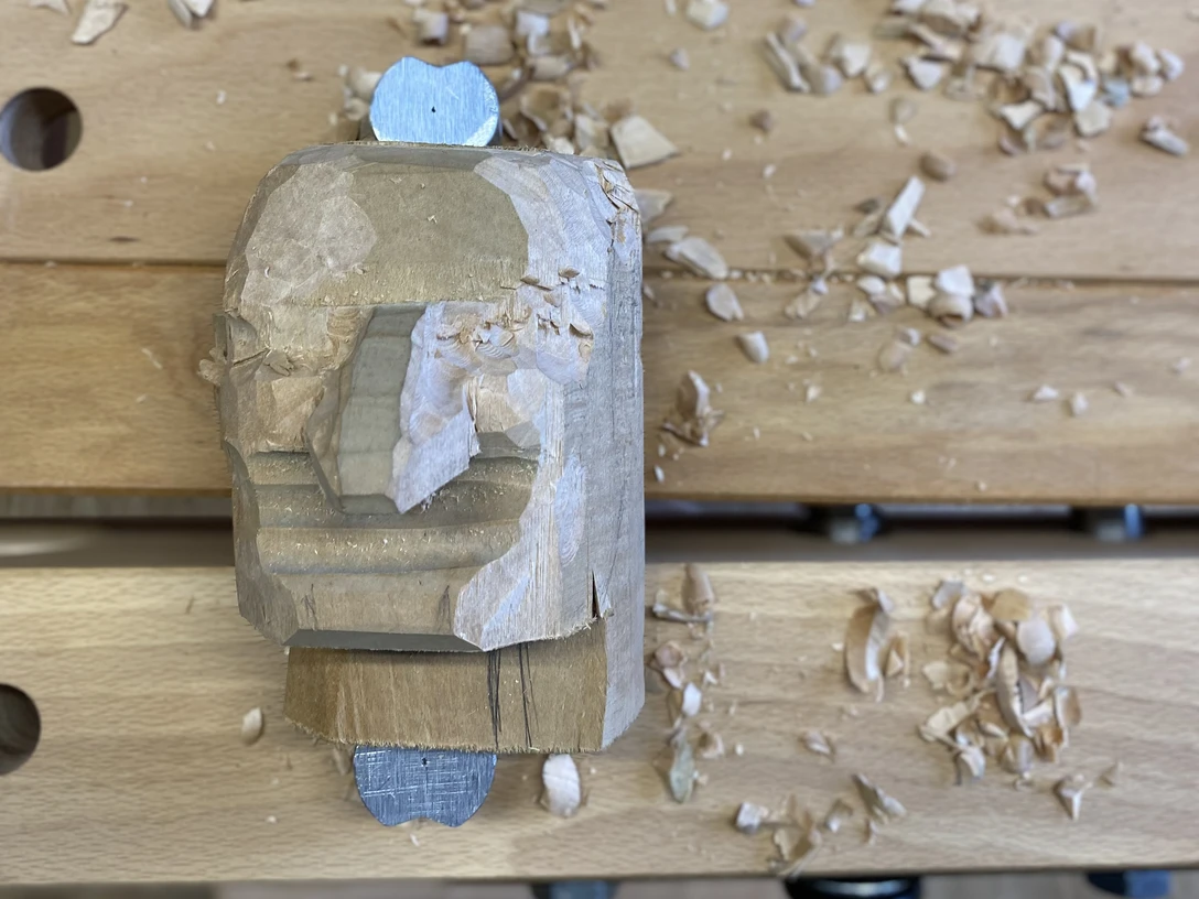 kasper-schnitzkopf-werkbank-schnitzbude-hohnstein-kasper-(c)-tvssw-madlen-rogge.jpeg Holzskulptur eines Gesichts in Arbeit auf einer Werkbank, umgeben von Holzspänen.