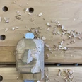 kasper-schnitzkopf-werkbank-schnitzbude-hohnstein-kasper-(c)-tvssw-madlen-rogge.jpeg Holzskulptur eines Gesichts in Arbeit auf einer Werkbank, umgeben von Holzspänen.