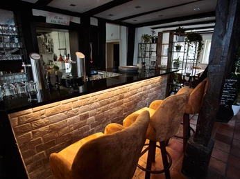 Prego_Rheda_5.jpg Moderne Bar mit gepolsterten Hockern, beleuchtetem Tresen und Gläsern vor rustikalem Hintergrund.