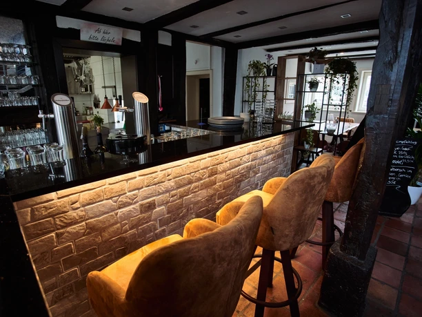 Prego_Rheda_5.jpg Moderne Bar mit gepolsterten Hockern, beleuchtetem Tresen und Gläsern vor rustikalem Hintergrund.