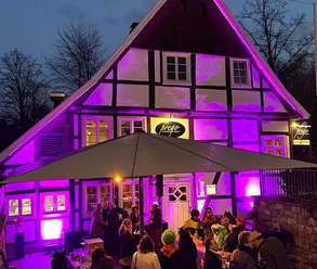 Fachwerkhaus bei Nacht, in lebendigem Lila angestrahlt, mit Besuchern unter einem großen Schirm.
