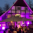 Prego_Rheda_1.jpg Fachwerkhaus bei Nacht, in lebendigem Lila angestrahlt, mit Besuchern unter einem großen Schirm.