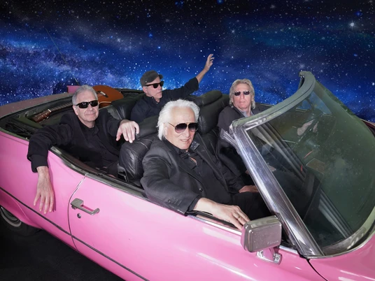 Hannes Schöner Band Vier ältere Männer in Sonnenbrillen fahren in einem pinken Cabrio durch eine Galaxie-Kulisse.Four older men in sunglasses drive through a galaxy backdrop in a pink convertible.