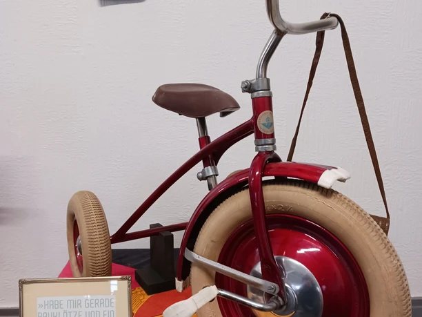 Sonderausstellung Ein rotes, nostalgisches Dreirad mit weißen Reifen steht auf einem bunten Tuch, umgeben von Spielklötzen.