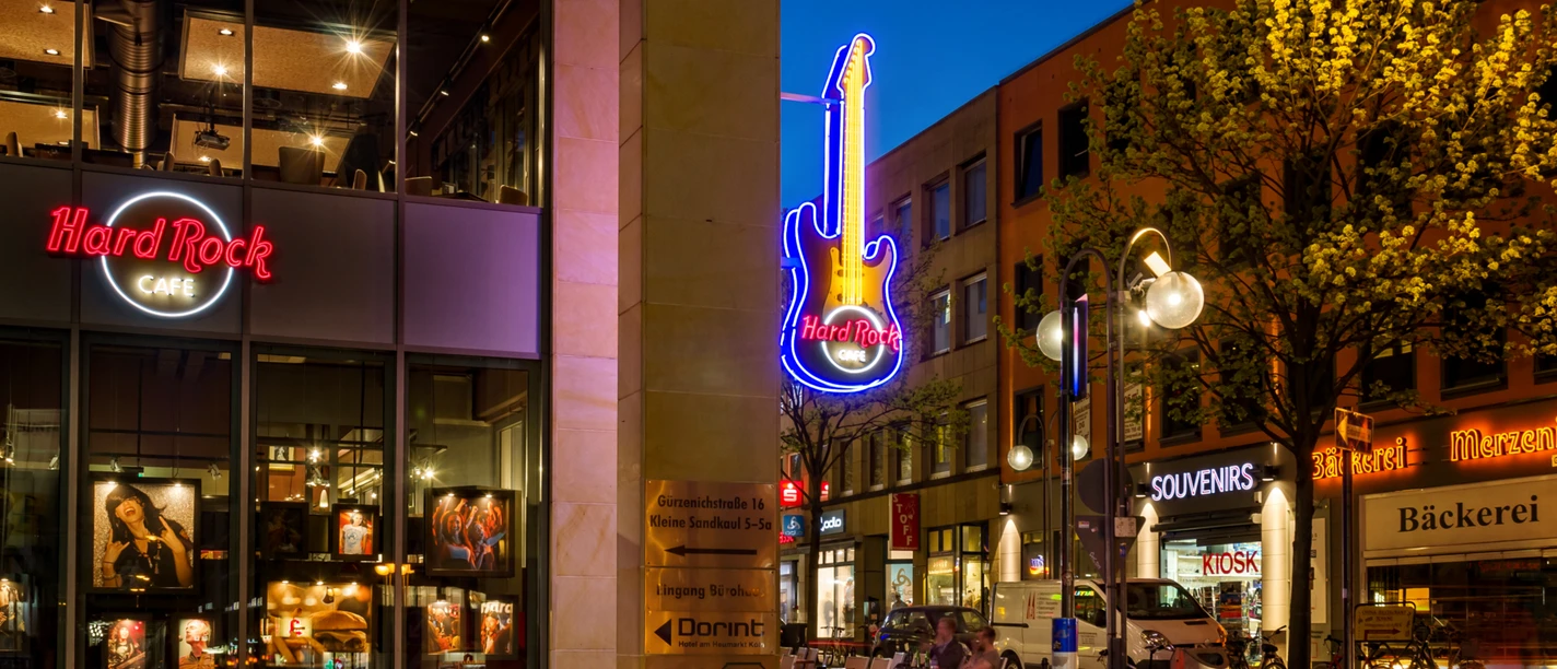 Exterior view of the Hard Rock Cafe with illuminated guitar and surrounding stores at night.</p> <p