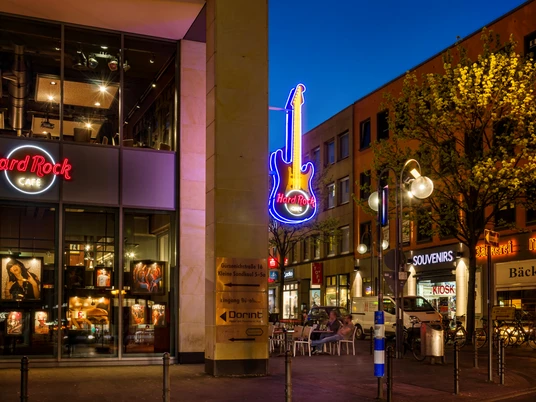 HRC Cologne_Exterior Außenansicht des Hard Rock Cafes mit beleuchteter Gitarre und umliegenden Geschäften in der Nacht.</p>Exterior view of the Hard Rock Cafe with illuminated guitar and surrounding stores at night.</p> <p