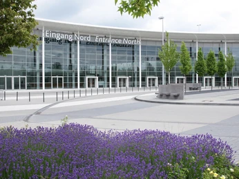 Congress-Centrum Koelnmesse Nord, North Halls entrance Glasgebäude mit Eingangsbereich, flankiert von Bäumen und lila Blumen im Vordergrund.</p>Glass building with entrance area, flanked by trees and purple flowers in the foreground.</p> <p