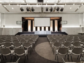 Congress-Centrum Koelnmesse Ost, Congress-Saal Moderne Konferenzhalle mit leeren Stuhlreihen und großer Bühne unter hellen Deckenleuchten.</p>Modern conference hall with empty rows of chairs and a large stage under bright ceiling lights.</p> <p