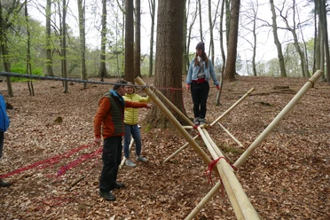 Brückenbau_kreativ.JPG Menschen balancieren auf einem Holzparcours im Wald unter Anleitung, umgeben von Laub auf dem Boden.</p>