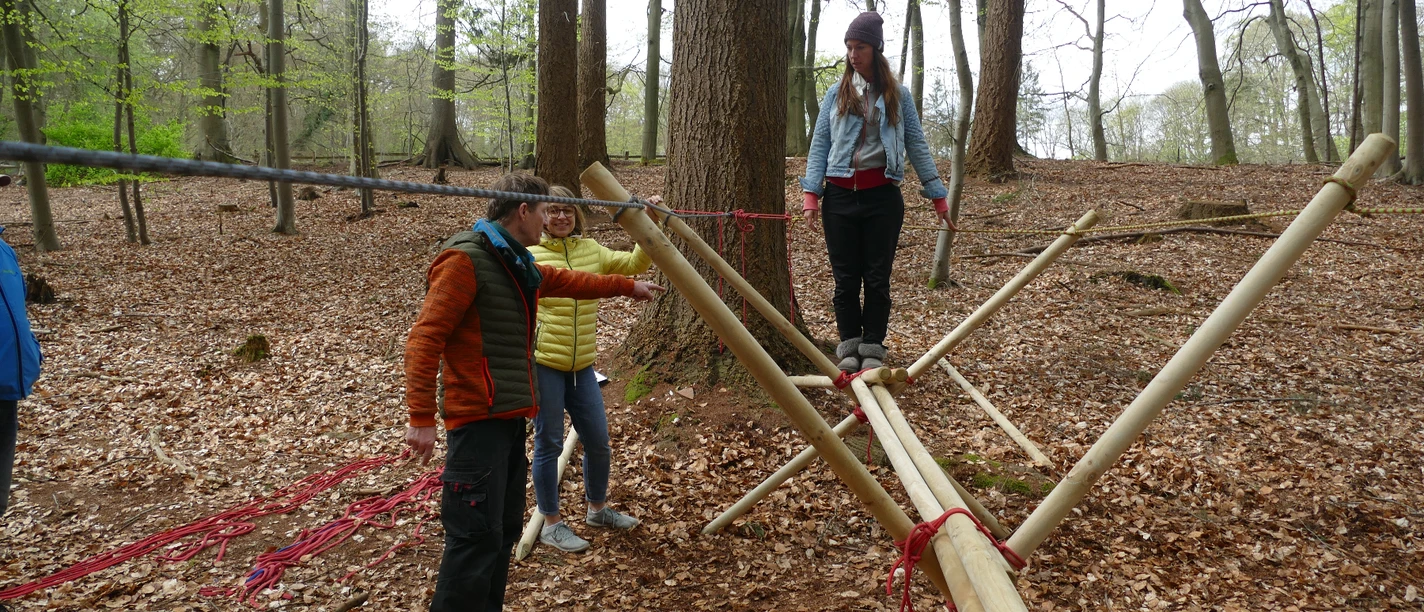 Brückenbau_kreativ.JPG Menschen balancieren auf einem Holzparcours im Wald unter Anleitung, umgeben von Laub auf dem Boden.</p>