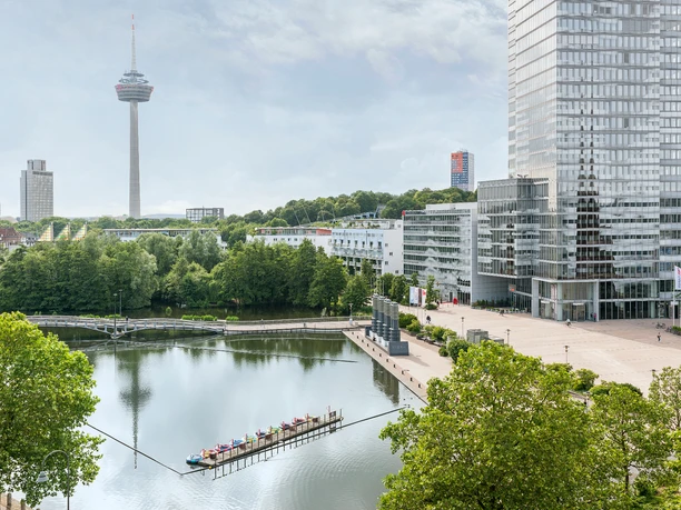 NH_collection-köln-mediapark Blick auf einen modernen Gebäudekomplex am See mit Turm im Hintergrund, umgeben von viel Grün.</p>