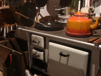 Küche mit historischem Herd, rostiger Teekanne und alten Pfannen, umgeben von Kochutensilien.