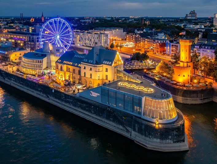 Schokoladenmuseum Gesamtansicht Blick auf beleuchtetes Riesenrad und Altstadt am Düsseldorfer Rheinufer bei Dämmerung.</p>View of the illuminated Ferris wheel and old town on the banks of the Rhine in Düsseldorf at dusk.</p> <p