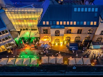 Vogelperspektive Terrasse Luftaufnahme eines beleuchteten Gebäudes mit Terrasse und Gästen unter Sonnenschirmen bei Nacht.</p>Aerial view of an illuminated building with terrace and guests under parasols at night.</p> <p