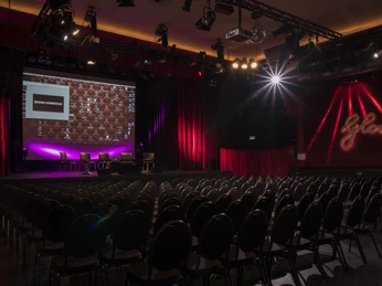 Gloria-Saal_bestuhlt_Sicht auf Bühne_quer Leerer Theatersaal mit Bühne und beleuchteten Vorhängen im Gloria Theater, rote Akzente im Hintergrund.Empty theater hall with stage and illuminated curtains in the Gloria Theater, red accents in the background.