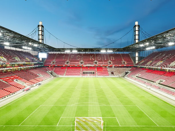 Innenbild RheinEnergie Stadion Interior view of an empty soccer stadium with green grass and rows of seats in red.</p> <p